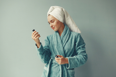 Woman in a blue bathrobe with a towel-wrapped head examining skincare bottles—symbolizing careful BHA product selection