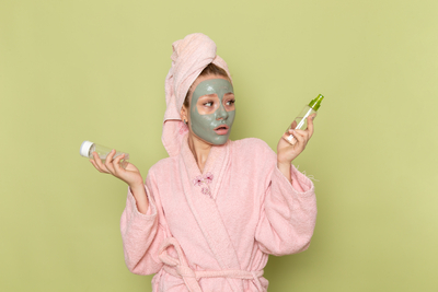 Confused woman in a pink robe with a green face mask comparing AHA vs BHA skincare bottles — choosing the best chemical exfoliant for her skin type.
