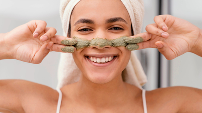Smiling woman applying a green clay mask to her nose, representing skincare tips to reduce the appearance of enlarged pores.
