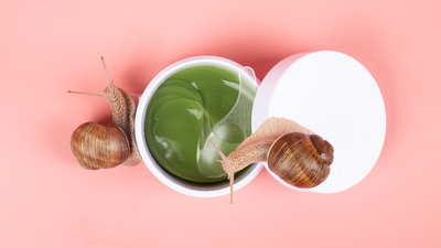 Two snails beside an open jar of green snail mucin gel on a pink background, representing K-beauty snail secretion filtrate for hydrated, glowy skin.