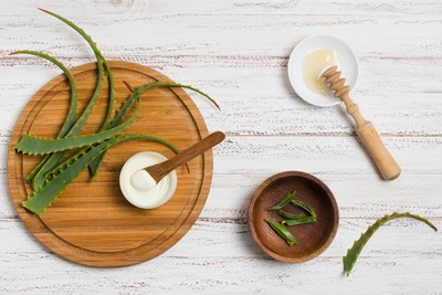 Fresh aloe vera leaves on a wooden board, a bowl of aloe gel, and a wooden honey dipper with honey on a white rustic table.