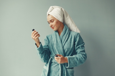  Woman in a blue bathrobe with a towel wrapped around her head, holding and examining two small skincare bottles against a plain background.