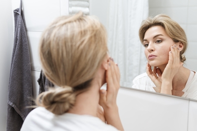 Woman examining acne-prone skin in the mirror, representing confusion between skin purging and breaking out after starting new skincare.